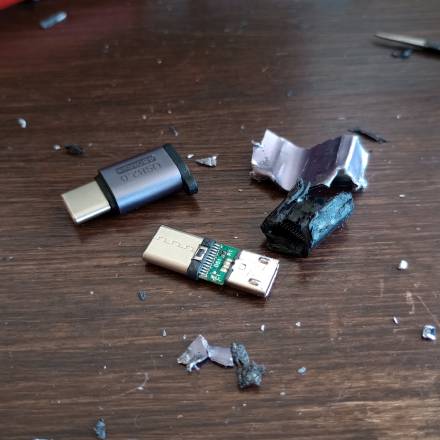 Überreste nach dem Zerlegen des Adapters: Plastik- und Aluminiumfetzen liegen verstreut um eine grüne Platine mit den beiden USB-Steckern. Ein noch neu wirkender Adapter liegt neben dem zerlegten.