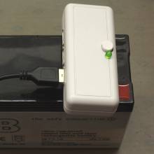 Die Powerbank in Benutzung. Ein kleines, rechteckiges graues Kästchen steckt auf den Kontaktfahnen eines Blei-Gel-Akkus. Auf der linken Seite ist ein USB-Kabel eingesteckt, oben sieht man einen grauen Einschalter neben einer grünen Betriebs-LED.