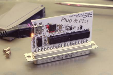 Fertig bestückte Platine mit großem Sub-D-Stecker (an der Platinenseite montiert), USB-Buchse (seitlich), Mikrocontroller, Quad-OPV, zwei DIP-Reed-Relais und einigen kleinen Bauteilen. Das leere Gehäuse liegt im Hintergrund.