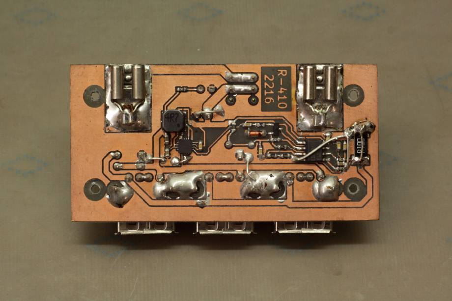 Die Unterseite der Platine, die keinen Lötstopplack hat. Auffällig sind größere Lötstellen für die Montagepins der USB-Buchsen und die Flachsteckkontakte (die einfach normale Crimp-Kontakte für Kabel sind). Das Schaltregler-IC ist ein winziges QFN nahe der Mitte der Platine, umgeben von der etwas größeren Spule und ein paar Kondensatoren. Die auf der Oberseite sitzenden USB-Buchsen schauen unten unter der Platine hervor.